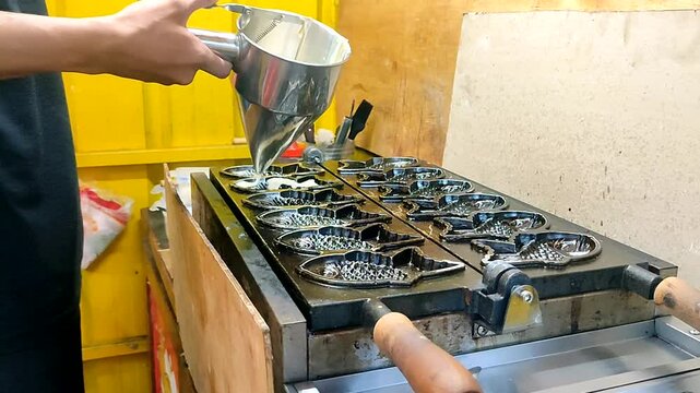 A man holds a stainless steel funnel filled with cake batter and pours it into a fish-shaped mold on a bungeo-ppang grill or waffle iron. Bungeo-ppang is a Korean  street food.