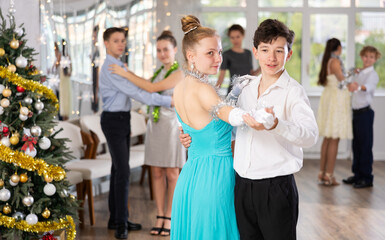 Enthusiastic adolescent dancers, girls and boys in elegant outfits practicing partner dance moves in studio adorned with Christmas decorations, under guidance of female teacher