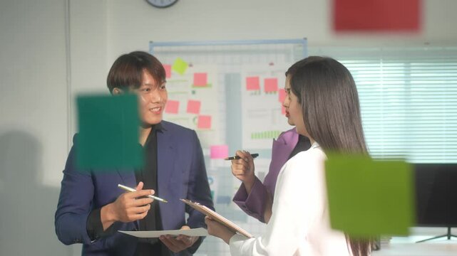 A man and a woman are talking in a room with a whiteboard. The man is holding a piece of paper and the woman is holding a clipboard. Scene is professional and focused