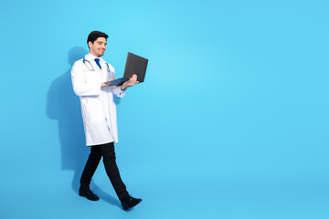 Young male doctor holding a laptop and walking on a vibrant blue background in a professional white...
