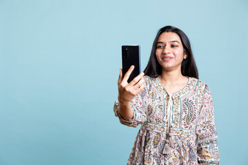 Fototapeta premium Indian woman attending a video call online from friends on her smartphone, stand against studio background. Portrait of a smiling girl having a fun discussion virtual meeting.