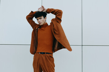 Young man striking a pose against a minimalist gray wall in a stylish brown outfit, showcasing...