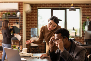 Male manager working with female employee, reviewing business documents and online data on laptop. Black man adjusting his glasses as asian businesswoman presents research statistics on digital device