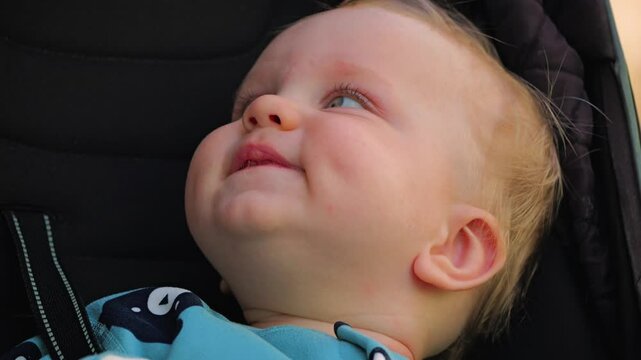 Close-up of the face of a cute baby sitting in a stroller. The baby has blue eyes, dimples on his cheeks and two bottom teeth. Happy baby boy on a walk