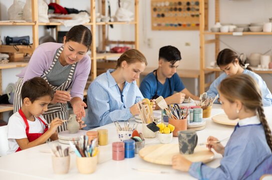 Teenagers and children at a ceramics workshop in an art studio. Woman teacher shows how to paint clay dishes with a brush
