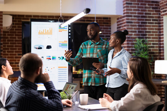 Black man and woman explaining business analytics to startup team, using led screen to illustrate trends and project plans during strategy meeting. Male employee with clipboard sharing project ideas.