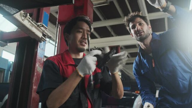 Two male mechanics cooperate, Latin and Asian workers fixing automotive wheels and maintaining car suspensions in garage station, and technician engineering service jobs in vehicle transport industry.