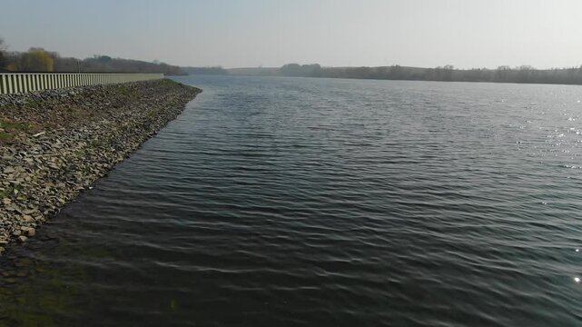 Calm lake view along rocky riprap embankment and distant trees under hazy sky, Dahlen, Germany. Aerial forward, low altitude, copy space