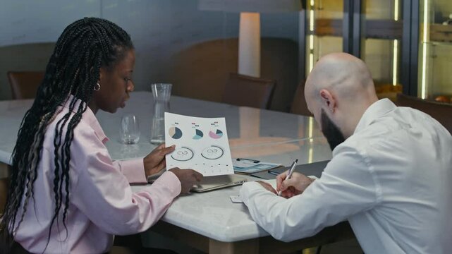 Medium shot of intercultural colleagues discussing financial chart when sitting at table in office space