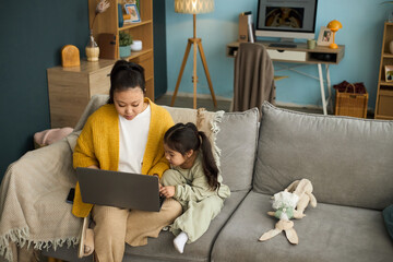 Mother and daughter sitting on cozy couch, engaging with laptop in warm living room