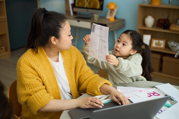 Mother assisting young daughter with schoolwork at table, surrounded by learning materials and books, they both focusing on document