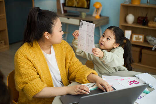 Mother assisting young daughter with schoolwork at table, surrounded by learning materials and books, they both focusing on document
