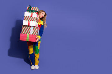 Young woman in colorful outfit holding a pile of stacked holiday gifts against a purple background