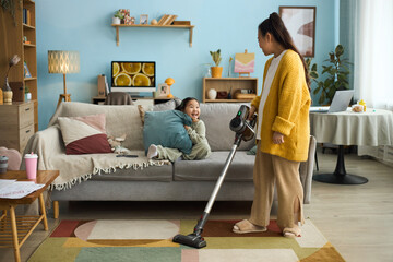 Woman vacuuming living room while child enjoying on sofa, cozy home environment with modern decor,...