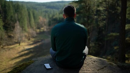 Man sits cross legged on clifftop edge looking out over forest landscape, practicing meditation or contemplation in the quiet of nature, concept of solitude and mental well being outdoors