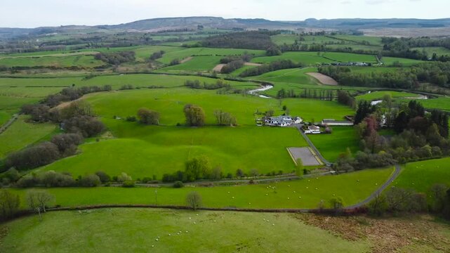 Aerial drone view of Scotland green grassy fields that has tall trees and small houses rooftops visible in front of bigger hills in the background during a cludy day. Roads and fences also visible.