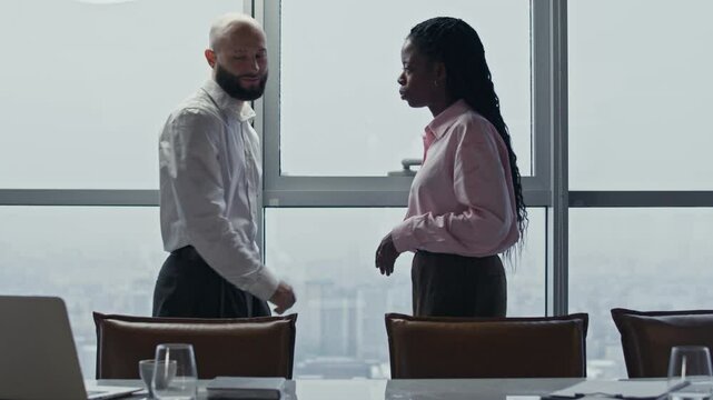 Side view of bicultural entrepreneurs in formalwear shaking hands by panoramic windows at office space