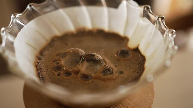 Dark roasted coffee is slowly dripping through a white paper filter placed in a clear glass v60 dripper, creating a mesmerizing display of bubbles and foam on the surface