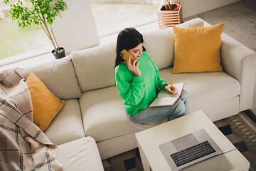 Young woman in green sweatshirt multitasking at home with notebook, phone, and laptop in a modern...