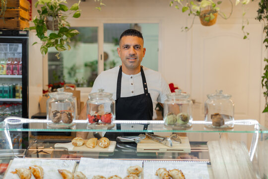Smiling man in a light shirt and apron stands behind the counter of a small food shop. Jars with cookies and empanadas are displayed. Hospitality and small business concept. Horizontal, copy-space.