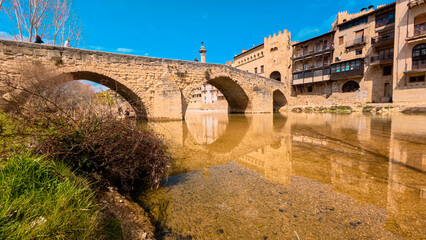 Fototapeta premium Stone bridge of Valderrobres over the Matarraña river seen from below, with the medieval old town in the background. Spring scene in the Matarraña region, Teruel, Spain. 