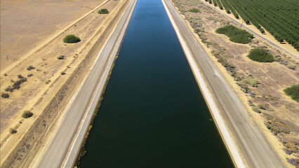 a vast irrigation canal bordered by dry, arid land on one side and lush green rows of trees on the...