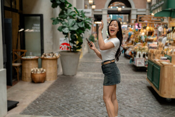 Young woman waving while holding smartphone in shopping mall