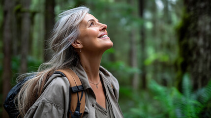 Naklejka premium A middle-aged woman in the forest. She looks up with a smile on her face. She has gray hair, brown eyes, and white teeth. She is wearing a khaki jacket and has a backpack on her shoulder.