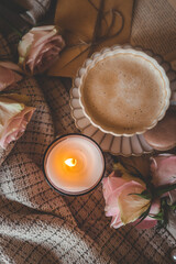 Top view of a cozy setting with a cup of coffee, macaron, pink roses, a lit candle, and an open book on soft textured fabric
