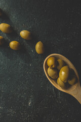 Top view of green olives in a wooden spoon with scattered olives on a dark background. Rustic and moody composition highlighting natural food textures