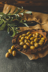 Rustic wooden bowl filled with green olives on burlap fabric with olive branch in background....