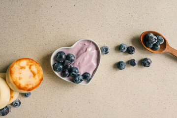 Two cottage cheese pancakes with blueberries and heart-shaped bowl of yogurt on light textured background, top view