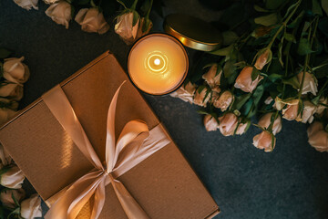 Top view of a glowing candle, beige roses, and a gift box tied with satin ribbon, arranged on a dark stone background in a cozy setting