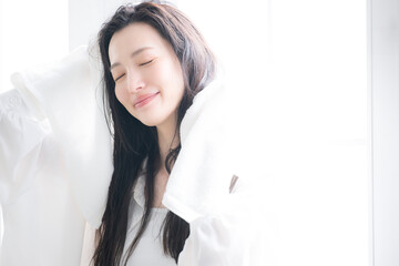 逆光の窓辺で朝やお風呂上りなどに濡れた髪を乾かすタオルドライやヘアケアする女性　目を閉じる