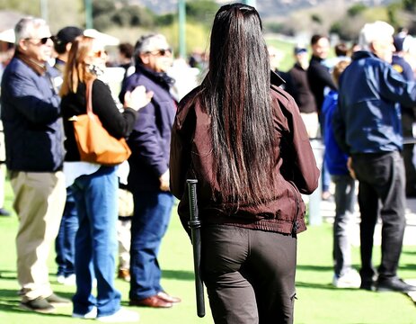 Vigilante de seguridad privada mujer con porra de espaldas protegiendo recinto al aire libre entre personas aficionadas de p&uacute;blico