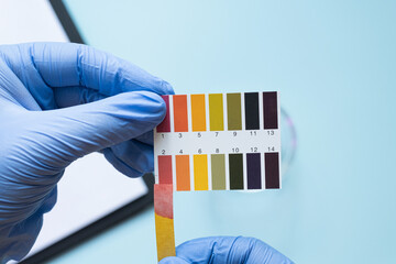 Hands holding ph test strip with color scale on blue background. Acidic pH environment.
