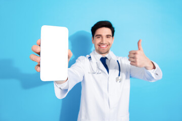 Smiling doctor presenting a smartphone with a white screen against a blue background while giving a thumbs up and wearing a white coat