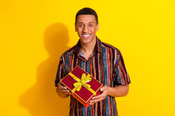 Smiling young man holding a gift box against a vibrant yellow background, showcasing happiness, leisure