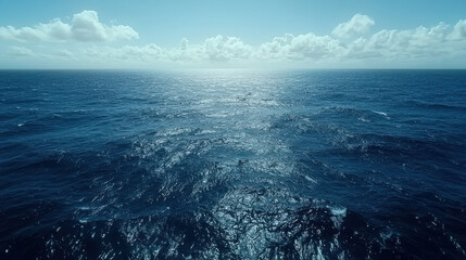 A breathtaking panoramic view of a vast, deep blue ocean surface reflecting sunlight under a partly cloudy sky on a tranquil day.