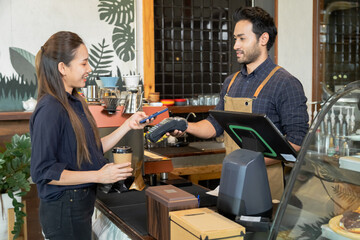 Young Indian baristas or cashier workers man holding a payment machine for scan, cashless technology and credit card payment concept. Customer using smartphone for payment to owner at cafe restaurant