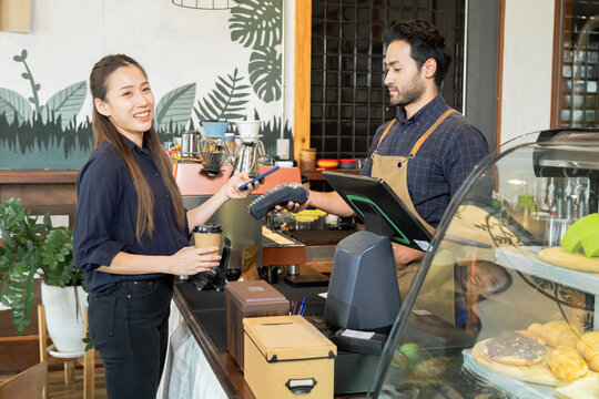 Young Indian baristas or cashier workers man holding a payment machine for scan, cashless technology and credit card payment concept. Customer using smartphone for payment to owner at cafe restaurant