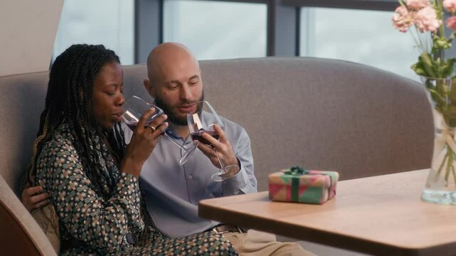 Pan shot of affectionate bicultural couple drinking red wine while having romantic dinner with present in restaurant