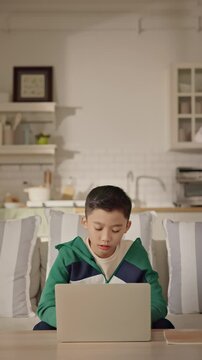 A young Asian boy sits comfortably at home using a laptop for gaming, studying, or browsing online. Modern childhood, digital education, balance of learning and leisure through technology. Gen Alpha.