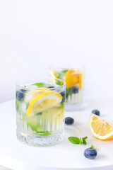 Chilled citrus drink with lemon slices, blueberries, mint, and ice in a glass and jug, on a white background.