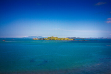 A stunning ocean view featuring a red ferry boat sailing near lush green islands in Auckland, New Zealand. The deep blue water contrasts beautifully with the vibrant landscape under a clear blue sky.