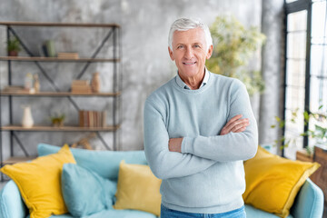 Señor mayor de 60 años con pelo blanco, de pie, con los brazos cruzados, vistiendo un jersey de punto azul pastel, con fondo desenfocado de Salón con sofá turquesa, cojines coloridos y estanteria