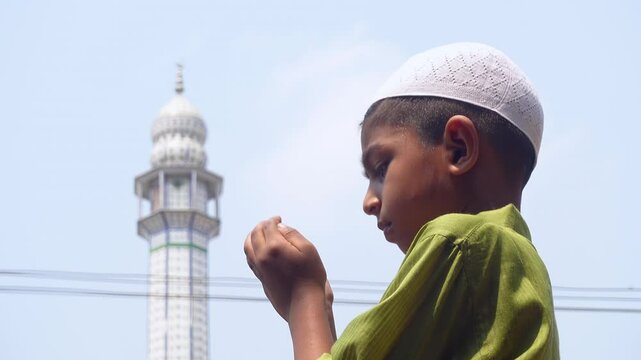 A Muslim boy stands in front of the minaret of a mosque and prays to God.