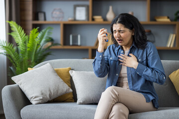 Young Indian woman suffering from asthma and allergies sitting on sofa at home, holding her hand on her chest and using inhaler