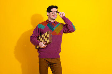 Smiling young man wearing glasses holding a chessboard against a vibrant yellow background,...