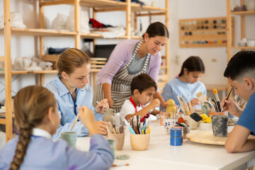 Children paint ceramics with brushes at the table in the creative studio. Teacher shows and helps children at the master class on ceramics